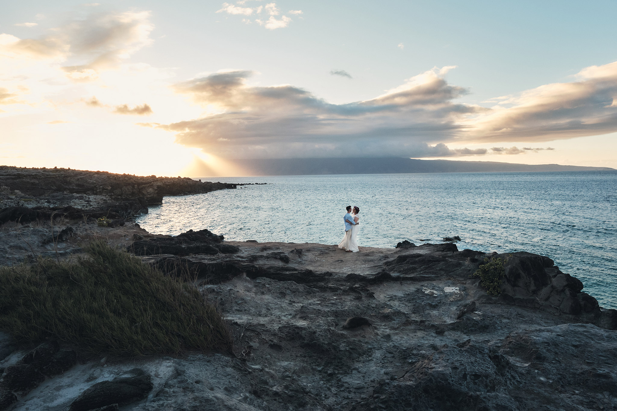 Eloping couple on the in an embrace on the side of a cliff overlooking the ocean at sunset.