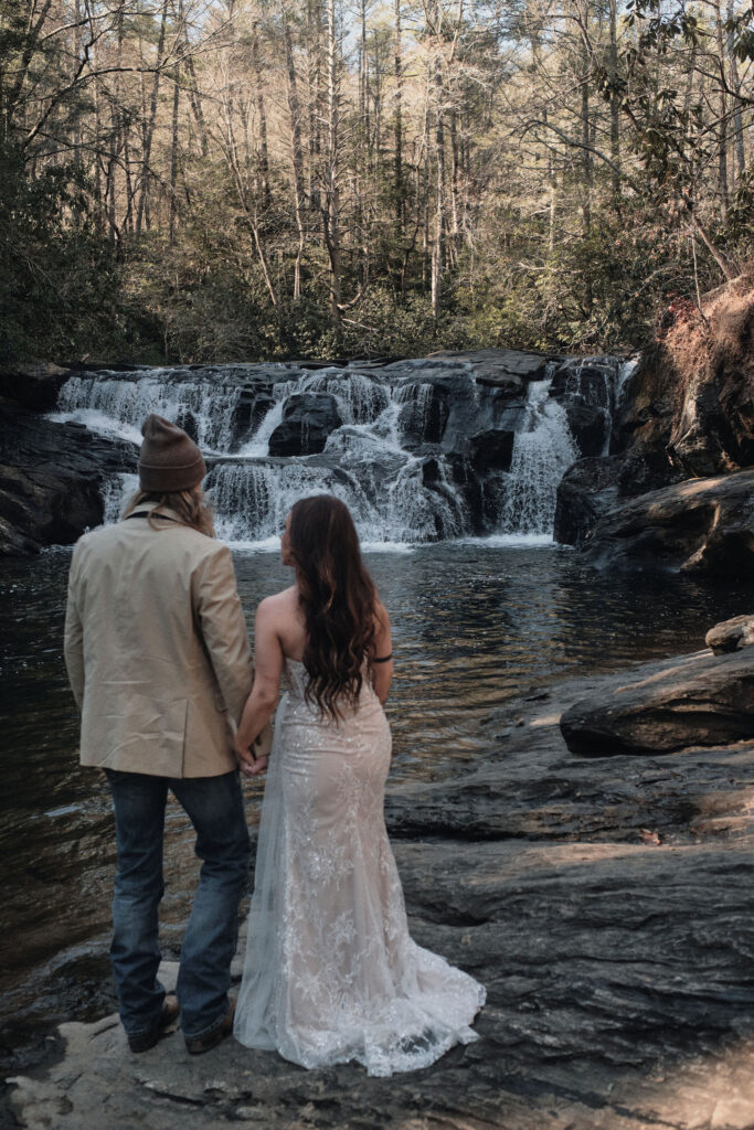 Eloping couple holding hands looking at a waterfall.